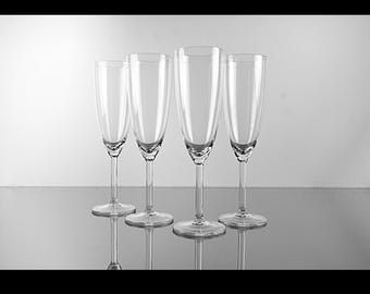 May include: Four clear champagne flutes of varying heights are arranged in a row. The glasses have long stems and wide bowls, reflecting the light. The background is a gradient of white to gray.