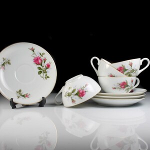 Peut inclure: Un ensemble de quatre tasses &agrave; th&eacute; et soucoupes blanches avec un motif floral de roses roses et de feuilles vertes. Les tasses sont empil&eacute;es les unes sur les autres et les soucoupes sont dispos&eacute;es autour d'elles.