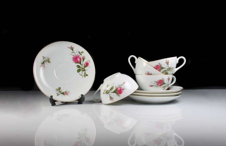 Peut inclure: Un ensemble de quatre tasses &agrave; th&eacute; et soucoupes blanches avec un motif floral de roses roses et de feuilles vertes. Les tasses sont empil&eacute;es les unes sur les autres et les soucoupes sont dispos&eacute;es autour d'elles.