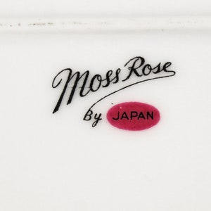 Puede incluir: Un plato de cer&aacute;mica blanco con texto en cursiva negra que dice "Moss Rose" y "By Japan" en un &oacute;valo rojo.