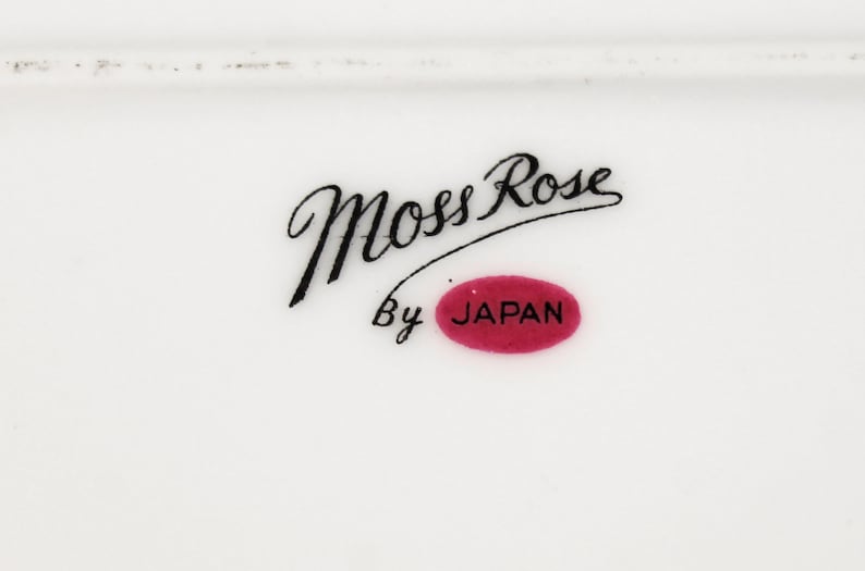 Puede incluir: Un plato de cer&aacute;mica blanco con texto en cursiva negra que dice "Moss Rose" y "By Japan" en un &oacute;valo rojo.