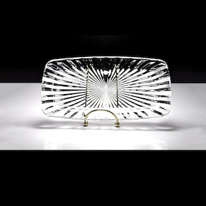 May include: A rectangular clear glass tray featuring a prominent sunburst pattern of radiating ridges. The tray is supported by a gold-toned metal stand with a curved base and two vertical prongs that hold the glass piece upright.