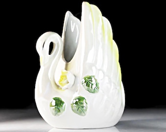 May include: White ceramic swan vase with green leaves and yellow flowers. The swan's wings are spread out and the vase is decorated with a floral design.