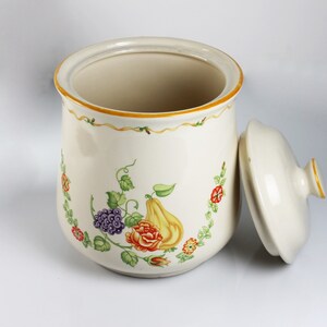 May include: A white ceramic canister with a floral design featuring a pear, grapes, and roses. The canister has a lid and is decorated with a yellow and green border.
