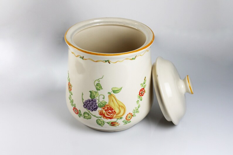 May include: A white ceramic canister with a floral design featuring a pear, grapes, and roses. The canister has a lid and is decorated with a yellow and green border.