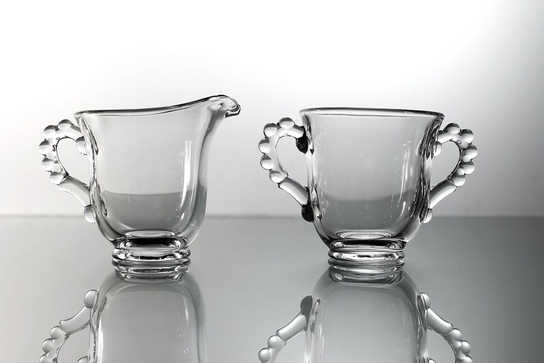 K&ouml;nnte beinhalten: Ein Set aus zwei klaren Glaskr&uuml;gen f&uuml;r Sahne und Zucker mit perlenbesetzten Griffen. Die Kr&uuml;ge sind leer und haben ein klassisches Design.