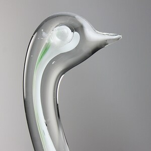 May include: Close-up of a clear glass swan sculpture with a white and green swirl design. The swan's head is tilted to the side, and its beak is slightly open.