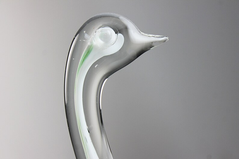 May include: Close-up of a clear glass swan sculpture with a white and green swirl design. The swan's head is tilted to the side, and its beak is slightly open.