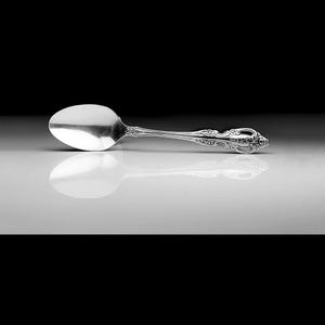 May include: A silver spoon with an ornate handle. The spoon is lying on a white surface with a reflection visible.