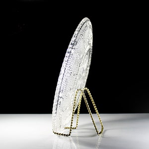Op de afbeelding: Een heldere glazen plaat met een decoratief ontwerp wordt getoond op een gouden metalen standaard. De plaat heeft een reli&euml;f, ingewikkeld patroon.