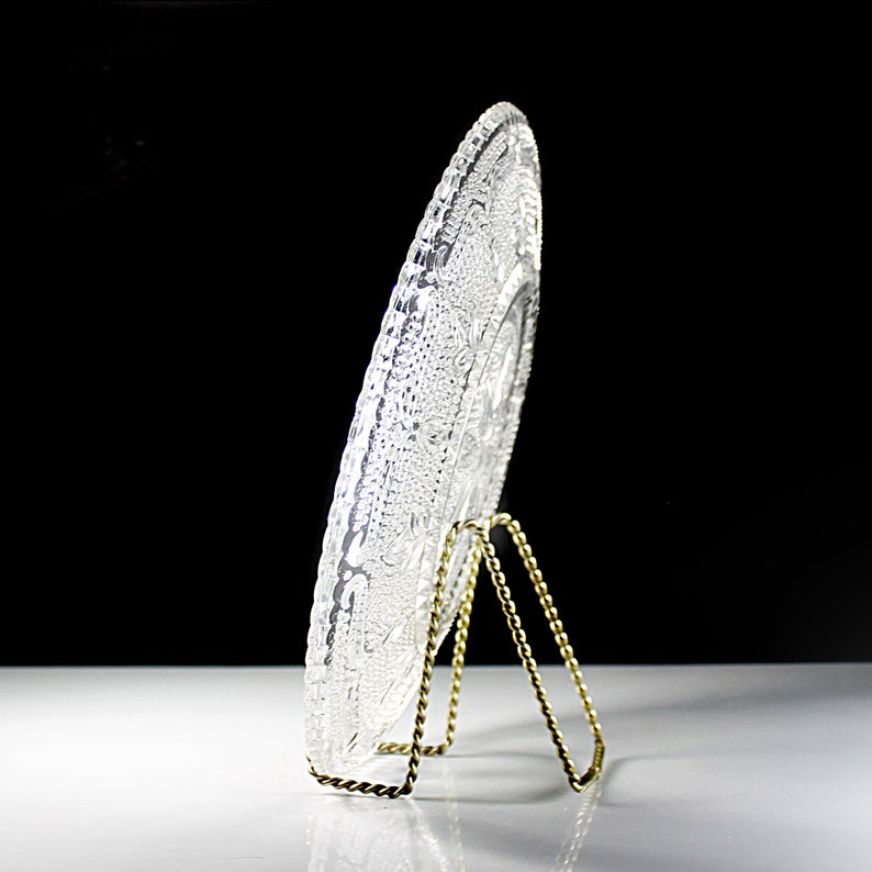 Op de afbeelding: Een heldere glazen plaat met een decoratief ontwerp wordt getoond op een gouden metalen standaard. De plaat heeft een reli&euml;f, ingewikkeld patroon.