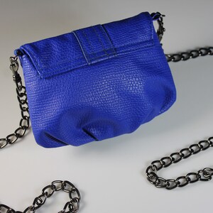 Pode incluir: Uma pequena bolsa azul cobalto com uma superf&iacute;cie texturizada e uma al&ccedil;a de corrente de metal. A bolsa tem um fecho de aba e um design simples. A corrente &eacute; drapeada sobre uma superf&iacute;cie branca.