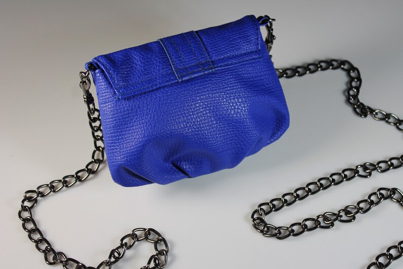 Pode incluir: Uma pequena bolsa azul cobalto com uma superf&iacute;cie texturizada e uma al&ccedil;a de corrente de metal. A bolsa tem um fecho de aba e um design simples. A corrente &eacute; drapeada sobre uma superf&iacute;cie branca.