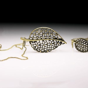 May include: A gold-toned necklace with a delicate leaf pendant. The pendant is made of intricate metalwork and has a lacy design. The necklace is on a chain with a lobster clasp.