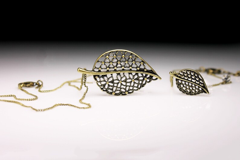 May include: A gold-toned necklace with a delicate leaf pendant. The pendant is made of intricate metalwork and has a lacy design. The necklace is on a chain with a lobster clasp.