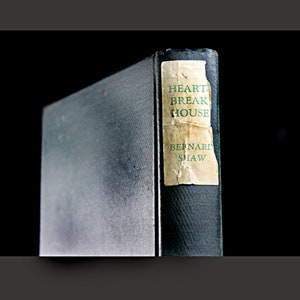 Puede incluir: Un libro antiguo de tapa dura titulado "Heartbreak House" de Bernard Shaw. El libro tiene una cubierta de color verde oscuro con una etiqueta amarillenta y descolorida.