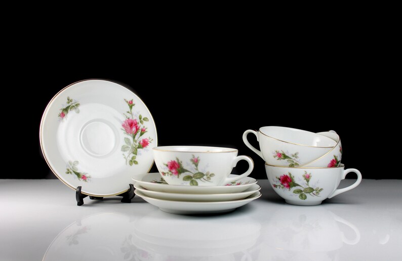 Peut inclure: Un ensemble de quatre tasses &agrave; th&eacute; et soucoupes blanches avec un motif floral repr&eacute;sentant des roses roses et des feuilles vertes. Les tasses et les soucoupes sont empil&eacute;es les unes sur les autres.