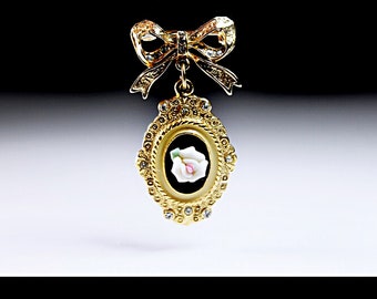 May include: A gold-tone brooch with a white cameo featuring a pink rose in a black oval frame. The brooch is attached to a gold-tone bow.