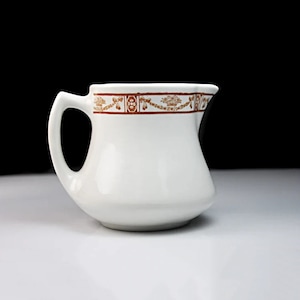 May include: White ceramic creamer with a brown decorative band around the top. The band features a repeating pattern of stylized flowers and crowns.