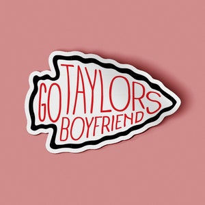 May include: A white sticker with a black outline in the shape of an arrowhead. The words "GO TAYLORS BOYFRIEND" are written in red inside the arrowhead.