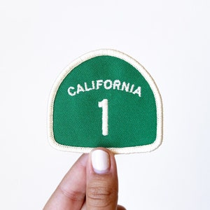 May include: A green and white California highway route patch with the number "1" in white on a green background. The patch is held by a hand.