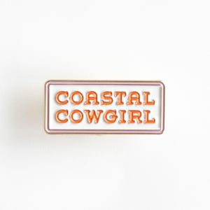 Peut inclure: Une épingle en émail blanc avec une bordure dorée et un contour rose. L'épingle dit "COASTAL COWGIRL" en lettres orange.