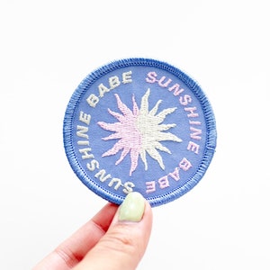 May include: A blue embroidered patch with a pink and white sun in the center. The text around the sun reads "Sunshine Babe Sunshine Babe".