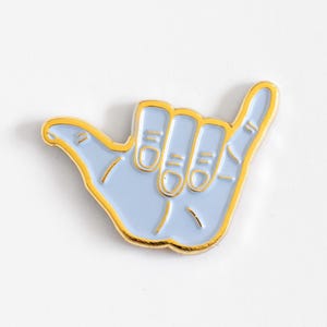 May include: A light blue enamel pin shaped like a hand making the shaka sign. The design features a gold-colored metal outline that defines the fingers, thumb, and palm, creating a clean and distinct border around the soft blue colored fill.