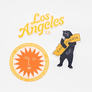 Peut inclure: Trois autocollants jaunes et orange avec un soleil, un ours tenant un contour de la Californie et le texte "Los Angeles CA".