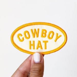 May include: A white oval patch with a yellow embroidered border. The text "COWBOY HAT" is embroidered in yellow on the patch.