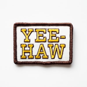 May include: Rectangular patch with the text "YEE-HAW" in yellow, outlined in brown, on a white background. The patch has a brown border with a stitched appearance. The design is centered on a white surface.