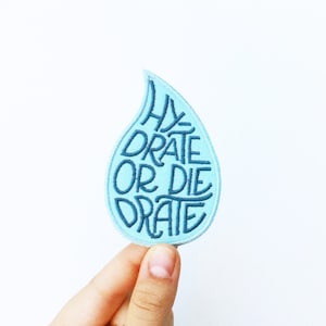 May include: A light blue, teardrop-shaped patch with the words "HYDRATE OR DIE DRATE" embroidered in dark blue. The patch is held against a white background.