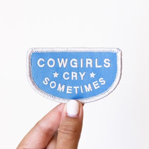 May include: A light blue fabric patch with white stitching and the text "COWGIRLS CRY SOMETIMES" with a star on either side of the word "CRY".