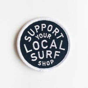 May include: A black and white embroidered patch with the text "Support Your Local Surf Shop".