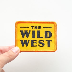 Peut inclure: Écusson brodé jaune avec du texte noir qui dit "THE WILD WEST" avec une bordure orange.