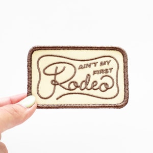 May include: A brown embroidered patch with a cream background. The patch features the text "Ain't My First Rodeo" in a cursive font.