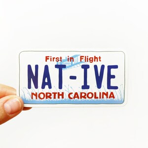 May include: A blue and white sticker with the text "First in Flight" and "NAT-IVE" in blue letters. The sticker also has the text "NORTH CAROLINA" in red letters.