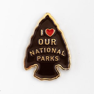 May include: An arrowhead-shaped enamel pin with a dark brown background and gold-coloured metal trim. The pin features a small red heart icon and the gold-coloured text I LOVE OUR NATIONAL PARKS arranged in four lines across the centre surface.