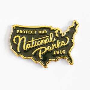 May include: A dark green enamel pin shaped like the map of the United States with gold-colored metal outlines and lettering. The text reads PROTECT OUR National Parks 1916 in a mix of block and cursive script fonts with gold-colored accents.