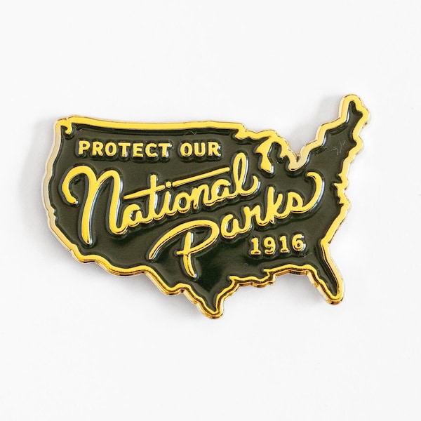 Pode incluir: Um broche de esmalte verde-escuro em formato de mapa dos Estados Unidos com contornos e letras em metal dourado. O texto diz PROTECT OUR National Parks 1916 em uma mistura de fontes de bloco e cursiva com detalhes em tom dourado.