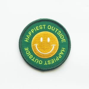 May include: Green embroidered patch with a yellow smiley face and the text "Happiest Outside" in a circle.