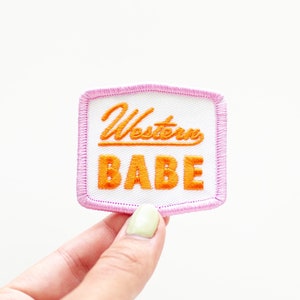 May include: A white patch with pink stitching around the edges. The patch has the text "Western Babe" embroidered in orange.