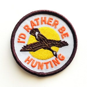 May include: Embroidered patch with the text "I'D RATHER BE HUNTING" in orange lettering. The patch features a brown bird in flight against a yellow circle, set on a white background, and framed by a dark brown border.