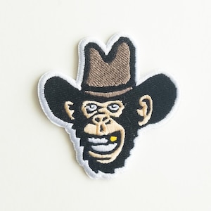 May include: Embroidered patch of a monkey wearing a brown cowboy hat with a gold tooth. The monkey is smiling with its mouth open.