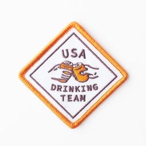 Peut inclure: Un écusson blanc en forme de diamant avec une bordure orange. L'écusson est brodé d'une image de deux mains tenant deux canettes de bière. Le texte "USA DRINKING TEAM" est brodé en marron sur l'écusson.