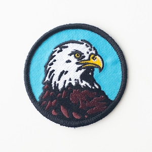 May include: An embroidered patch featuring a bald eagle's head, with a blue background, white feathers and a yellow beak.