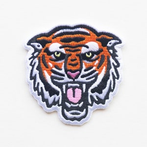 May include: A close-up of an embroidered iron-on patch featuring a tiger's face with an open mouth and a pink tongue. The tiger has orange and black stripes and black eyes.