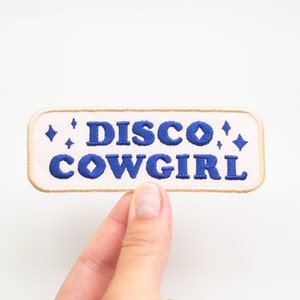 May include: A white embroidered patch with blue text that reads "Disco Cowgirl" with small blue stars on either side.