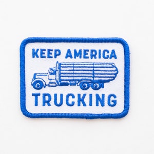 May include: A rectangular patch with a white background and a blue border. The patch features a blue illustration of a semi-truck carrying logs, with the words "KEEP AMERICA TRUCKING" in blue block letters above and below the truck image.