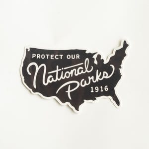 May include: A black and white sticker in the shape of the United States. The sticker reads "Protect Our National Parks 1916" in a mix of block and script lettering. The sticker is on a white background.